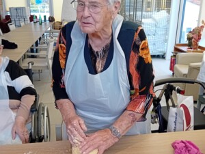 Eine Seniorin trägt eine Schürze und knetet einen großen Teigklumpen auf dem Tisch. Daneben stehen frische Eier und Backutensilien.