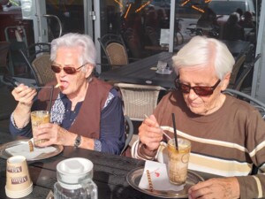 Zwei ältere Bewohnerinnen sitzen an einem Tisch im Außenbereich eines Cafés und essen Eis aus hohen Gläsern. Beide tragen Sonnenbrillen, auf dem Tisch stehen Servietten, Becher und eine Wasserflasche.