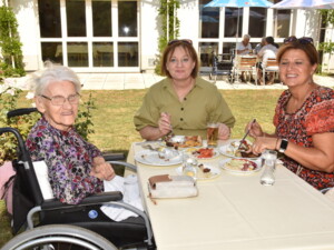 Eine Seniorin im Rollstuhl sitzt mit zwei Angehörigen an einem Gartentisch. Auf den Tellern liegen verschiedene Grillgerichte und Salate, im Hintergrund sind weitere Gäste im Garten zu sehen.