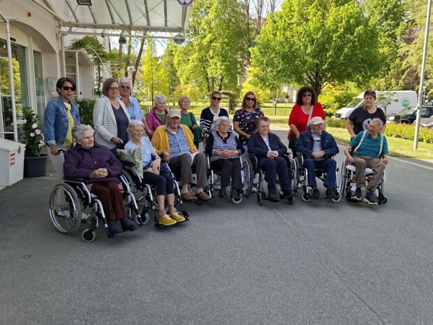 Eine Gruppe älterer Menschen, viele davon im Rollstuhl, sitzt und steht vor einem Gebäude im Freien. Betreuungspersonen begleiten sie. Im Hintergrund sind Bäume und ein sonniger Himmel zu sehen. Die Gruppe wirkt bereit für einen gemeinsamen Ausflug.