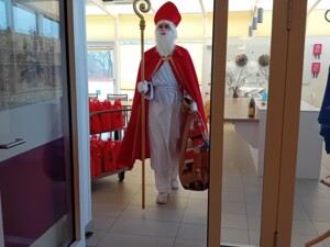 Der Nikolaus mit rotem Mantel und Mitra betritt durch eine Glastür einen Wohnbereich. In der Hand trägt er einen Bischofsstab, hinter ihm steht ein Wagen voller roter Geschenksäckchen.