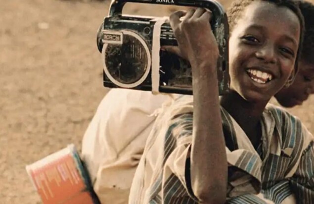 Symbolbild lächelndes afrikanisches Kind mit tragbarem Radio auf der Schulter im Vordergrund. Kind im Hintergrund sitzend.