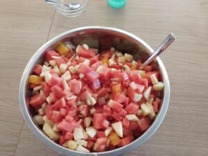 Eine große Metallschüssel ist mit gewürfeltem Obst gefüllt, darunter Wassermelone, Apfelstücke und gelbe Fruchtstücke. Ein Löffel steckt in der Schüssel.