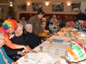 Eine als Clown verkleidete Person reicht einer sitzenden Seniorin ein Würstel. Weitere Bewohner:innen sitzen am Tisch mit Getränken und Gebäck. Im Hintergrund sind weitere Gäste und Mitarbeitende zu sehen. Bunte Dekorationen und Faschingshüte prägen das Bild.