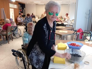 Eine Seniorin mit grünen Augenpads steht lächelnd vor einer Snackstation mit geschnittenem Obst wie Melone, Blaubeeren und Erdbeeren. Im Hintergrund sitzen weitere Bewohner:innen in einem freundlichen Raum.
