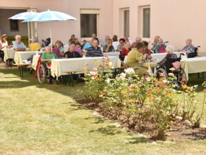 Weitwinkelaufnahme vom Garten mit mehreren gedeckten Tischen. Bewohner:innen und Gäste sitzen im Schatten oder unter Sonnenschirmen, essen und plaudern. Im Vordergrund blühen Rosenbeete.