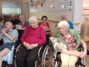 Mehrere ältere Bewohner:innen sitzen im Eingangsbereich des Hauses, einige in Rollstühlen. Hinter ihnen hängen ein Willkommensschild und Wegweiser zu den Wohnbereichen.