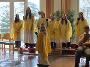 Mehrere Kinder in gelben Umhängen und mit goldenen Sternen als Kopfschmuck singen auf einer Bühne. Eine weitere Person steht davor und singt mit.