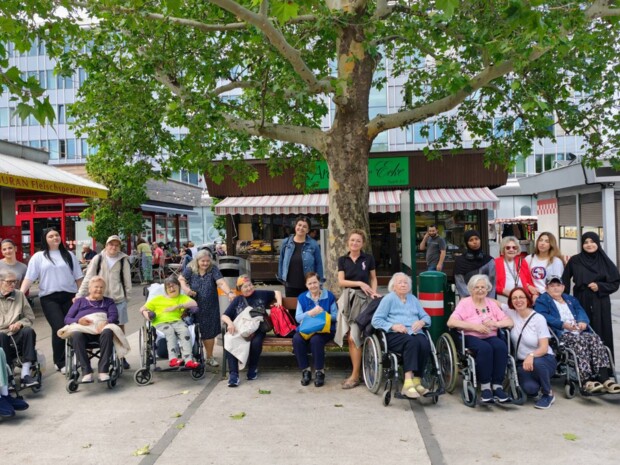 Eine große Gruppe von Menschen, darunter Senior:innen in Rollstühlen, Betreuungspersonen und junge Teilnehmer:innen von AusbildungsFit, posieren gemeinsam für ein Gruppenfoto unter einem großen Baum am Meidlinger Markt. Im Hintergrund sind Marktstände, ein Geschäft mit Fleischspezialitäten und ein Café zu sehen.