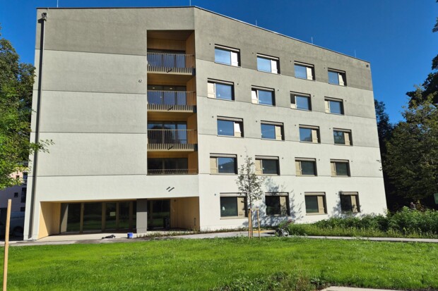 Ein fünfgeschossiges, modernes Wohngebäude mit vielen Fenstern, Balkonen und einer hellen Fassade. Es steht in einem gepflegten grünen Außenbereich mit Bäumen und Rasen.