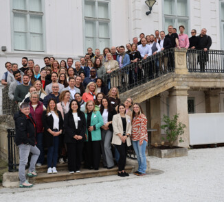 Große Gruppe von Teilnehmenden steht auf einer Treppe vor einem Gebäude und blickt in die Kamera für ein Erinnerungsfoto.