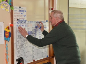 Ein älterer Mann steht vor einer großen Tafel im Eingangsbereich und schreibt mit einem Stift Wetterdaten auf. Auf der Tafel sind Datum, Temperaturen und weitere Messwerte notiert. Neben der Tafel hängt eine bunte Faschingsdekoration.