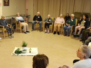 Lehrerin sitzend im Stuhlkreis mit SeniorInnen fröhlich sitzend, den Klängen der Intsrumente lauschend im Festsaal des Hauses.