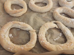 Mehrere goldbraun gebackene Fastenbeugel liegen auf Backpapier auf einem Backblech. Die ringförmigen Gebäckstücke sind unregelmäßig geformt und mit groben Salzkörnern bestreut.
