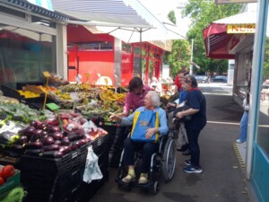 Mehrere Senior:innen, begleitet von Betreuungspersonen, betrachten das frische Obst- und Gemüseangebot eines Marktstandes am Meidlinger Markt. Eine Person im Rollstuhl zeigt auf das Angebot, während andere neben ihr stehen und mit ihr sprechen. Der Stand ist bunt bestückt mit Auberginen, Bananen, Tomaten und weiteren Produkten.