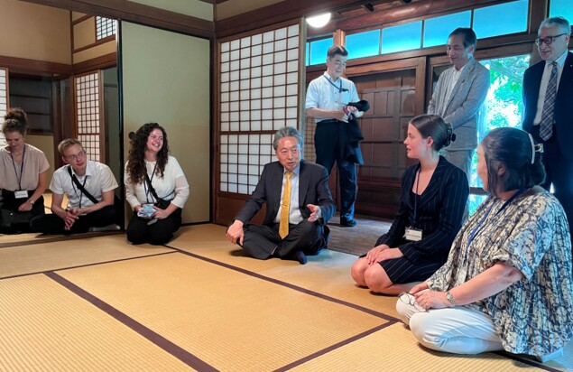 In einem traditionell japanisch eingerichtetem Haus knien mehrere Personen auf Tatami-Matten. Einige sind jung und europäisch, einige älter und japanisch. Alle sind formell gekleidet. Im Gang hinter dem Zimmer sind neben den offenen Papier-Schiebetüren einige weitere stehende Personen zu sehen.