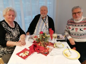 Drei Bewohner:innen sitzen an einem Tisch mit weihnachtlicher Dekoration, Getränken und kleinen Speisen. Sie blicken in die Kamera und wirken entspannt. Die Tischdeko besteht aus Tannenzweigen und roten Elementen, was eine festliche Stimmung erzeugt.