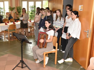 Eine junge Frau spielt auf der Steirischen Harmonika, daneben steht eine Gruppe Schüler:innen mit Liedtexten. Eine Lehrkraft steht im Hintergrund.