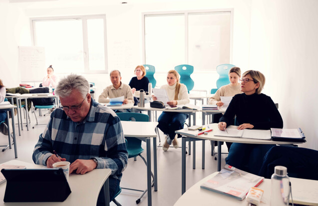Mehrere Kursteilnehmende sitzen in einem hellen Unterrichtsraum an einzelnen Tischen. Einige lesen oder halten Arbeitsblätter, eine Person im Vordergrund schreibt neben einem Tablet Notizen. Foto: www.inframe.at