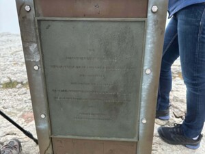Nahaufnahme der Gedenktafel am Sockel des ÖJAB-Gipfelkreuzes. Die Inschrift erinnert an das Jahr 1985 und betont den Einsatz für Freiheit, Zusammenarbeit und Opferbereitschaft. Einige Personen stehen im Hintergrund.