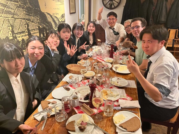 Zehn Personen sitzen an einem reich belegten, langen Holztisch und blicken in die Kamera. Die jungen Menschen sind formell gekleidet und essen teilweise noch. Manche strecken ihre Hände, Gläser oder ihr Besteck in die Höhe. Auf dem Tisch sind halbgefüllte Wein- und Wassergläser und Teller mit Resten von Brot, Käse, Aufstrichen und Braten zu sehen. Im Hintergrund sind rustikale Einrichtungsgegenstände wie ein Wagenrad oder ein altes Bild. Die Stimmung ist gesellig bis ausgelassen.