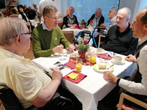 Bewohner:innen und Angehörige sitzen an einem Tisch, trinken Kaffee und Saft und unterhalten sich. Auf dem Tisch stehen Kuchenstücke, Tassen und Gläser. Die Personen sind einander zugewandt und wirken in angeregtem Gespräch. Die Szene vermittelt Gemeinschaft und Wohlbefinden.