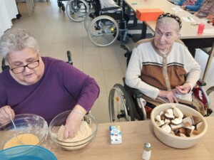 Zwei ältere Personen sitzen an einem Tisch und panieren gemeinsam frische Pilzstücke. Im Hintergrund sind weitere Bewohner:innen am Tisch beschäftigt.