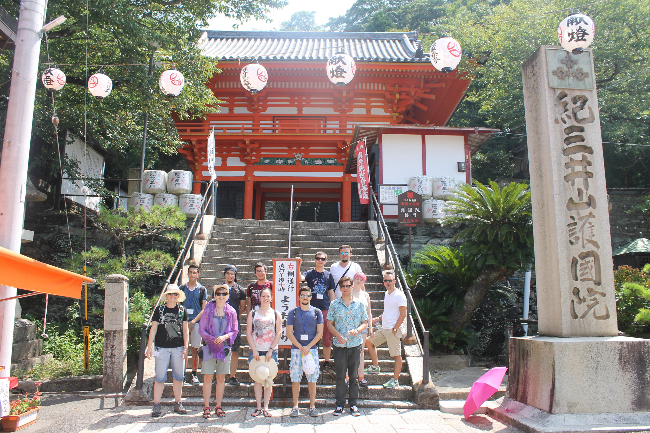 Eine Gruppe von zwölf Personen steht vor einem roten Tempeltor in der Präfektur Wakayama, Japan. Über der Treppe hängen Lampions, daneben stehen große Sakefässer und Steinlaternen. Im Vordergrund sind ein großer Stein mit japanischer Inschrift und eine pinke zusammengeklappte Schirm zu sehen. Die Gruppe nimmt im Rahmen einer von Kai-You-Kai organisierten Rundreise teil, die kulturelle Höhepunkte wie den heiligen Berg Kōya-san, den Pilgerweg Kumano Kodō sowie Workshops zu Kalligrafie und traditioneller Papierherstellung umfasst.