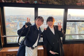 Zwei junge Japanerinnen stehen in einer verglasten Aussichtsgondel mit Blick über eine weitläufige Stadt bei warmem Abendlicht. Beide tragen dunkle Mäntel, lächeln in die Kamera und heben jeweils eine Hand zum Victory-Zeichen. Die linke Person hält zusätzlich eine Kamera, während im Hintergrund Dächer, Gebäude und Teile des Riesenrads bzw. der Gondelkonstruktion sichtbar sind.