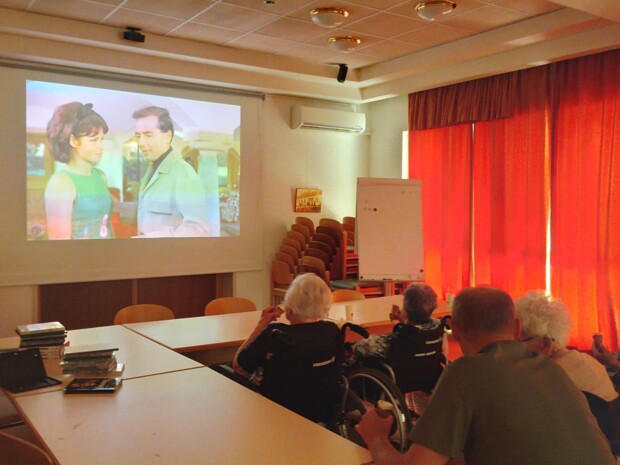 Bewohner:innen sitzen in einem Aufenthaltsraum vor einer großen Leinwand, auf der ein Heimatfilm gezeigt wird. Neben der Leinwand stehen gestapelte DVDs, rote Vorhänge verdunkeln den Raum.