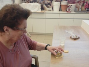 Eine ältere Frau sitzt an einem Tisch und sticht mit einer Form Keksteig aus. Neben ihr liegt ein Becher und eine offene Keksverpackung.