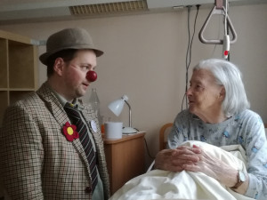 Clown sitzend neben Bewohnerin im Bett liegend.