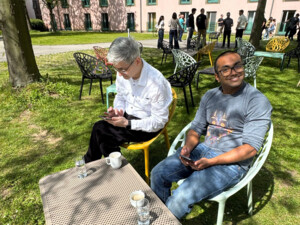 Zwei Personen sitzen auf bunten Stühlen im Freien mit Kaffeetassen und Handys, während im Hintergrund weitere Gruppen im Grünen stehen und sich unterhalten.