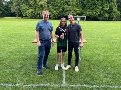 Drei Personen posieren gemeinsam auf einer Wiese, eine trägt eine Medaille. Die ausgezeichnete Person hält eine Getränkedose und trägt ein schwarzes Trikot.