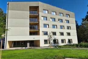 Ein fünfgeschossiges, modernes Wohngebäude mit vielen Fenstern, Balkonen und einer hellen Fassade. Es steht in einem gepflegten grünen Außenbereich mit Bäumen und Rasen.