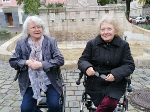 Zwei ältere Bewohner:innen sitzen mit Rollatoren vor einem steinernen Brunnen in einem ruhigen Stadtbereich. Hinter ihnen steht eine Statue, umgeben von Bäumen mit herbstlich gefärbten Blättern. Beide Personen blicken entspannt in die Kamera und halten persönliche Gegenstände in den Händen.