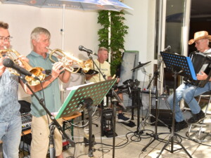 Vier Musiker spielen gemeinsam: Zwei mit Trompeten, einer am Schlagzeug und einer mit Akkordeon. Vor ihnen stehen Mikrofone und Notenständer.