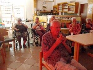 Bewohner:innen sitzen verteilt im Aufenthaltsraum und genießen Eis und Getränke. Im Vordergrund sitzt eine Person mit Eis, im Hintergrund sind weitere Bewohner:innen, Rollstühle, Tische und eine Bar zu sehen.