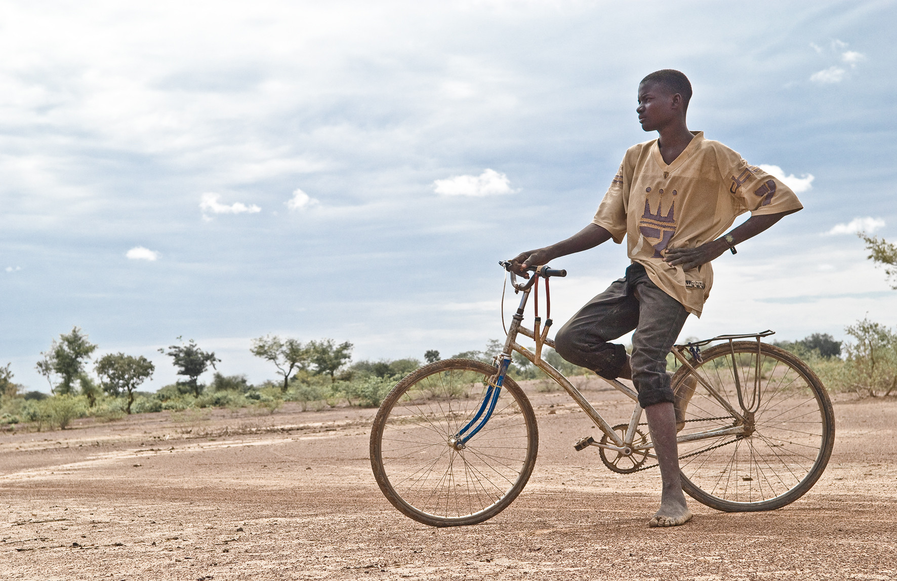 Junger Bewohner Burkina Fasos auf einem Fahrrad sitzend blickt nachdenklich in afrikanische Landschaft.
