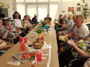 In einem hellen Gemeinschaftsraum sitzen Bewohner:innen rund um einen langen Tisch. Gemeinsam mit Schüler:innen der SOB Güssing bewegen sie farbige Tücher im Sitzen. Auf dem Tisch stehen Blumen, Becher und Klangschalen.