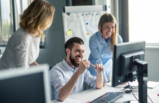 Ein Mann sitzt bei Tisch, lacht und schaut auf den Computer. Links und rechts von ihm sind zwei Frauen, die ebenfalls lachen und auf den Computerbildschirm schauen. 