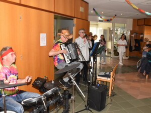 Drei Musiker spielen in der Eingangshalle eines Pflegeheims Faschingsmusik: Eine Person sitzt am Schlagzeug mit buntem Stirnband und Batikshirt, eine weitere spielt Akkordeon mit Cowboyhut, daneben bläst ein Musiker Trompete. Vor ihnen stehen Mikrofone und Lautsprecher. Im Hintergrund sind Mitarbeitende und Bewohner:innen zu sehen, die zuhören.