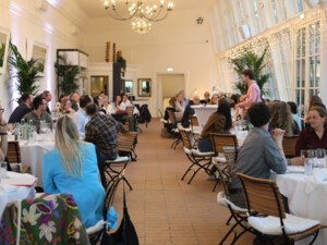 Großer Raum mit gedeckten Tischen, an denen viele Teilnehmende sitzen und Gesprächen folgen; eine Person spricht vor der Gruppe.