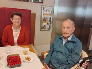 Zwei ältere Bewohner:innen sitzen an einem gedeckten Tisch mit Getränken, Kirschen und Knabbereien. Im Hintergrund sind ein Aufzug und Informationsmaterial zu sehen.