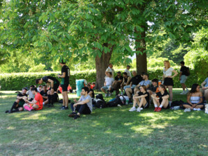 Viele junge Erwachsene sitzen und stehen unter großen Bäumen auf einer Wiese. Einige tragen Fußballtrikots, andere Alltagskleidung; Taschen und Rucksäcke liegen am Boden.