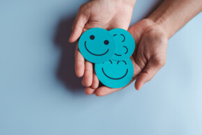 Symbolbild. Zwei geöffnete Hände halten drei türkisfarbene runde Karten mit verschiedenen Smileys, die Freude und positive Emotionen ausdrücken. Der Hintergrund ist einfarbig hellblau.