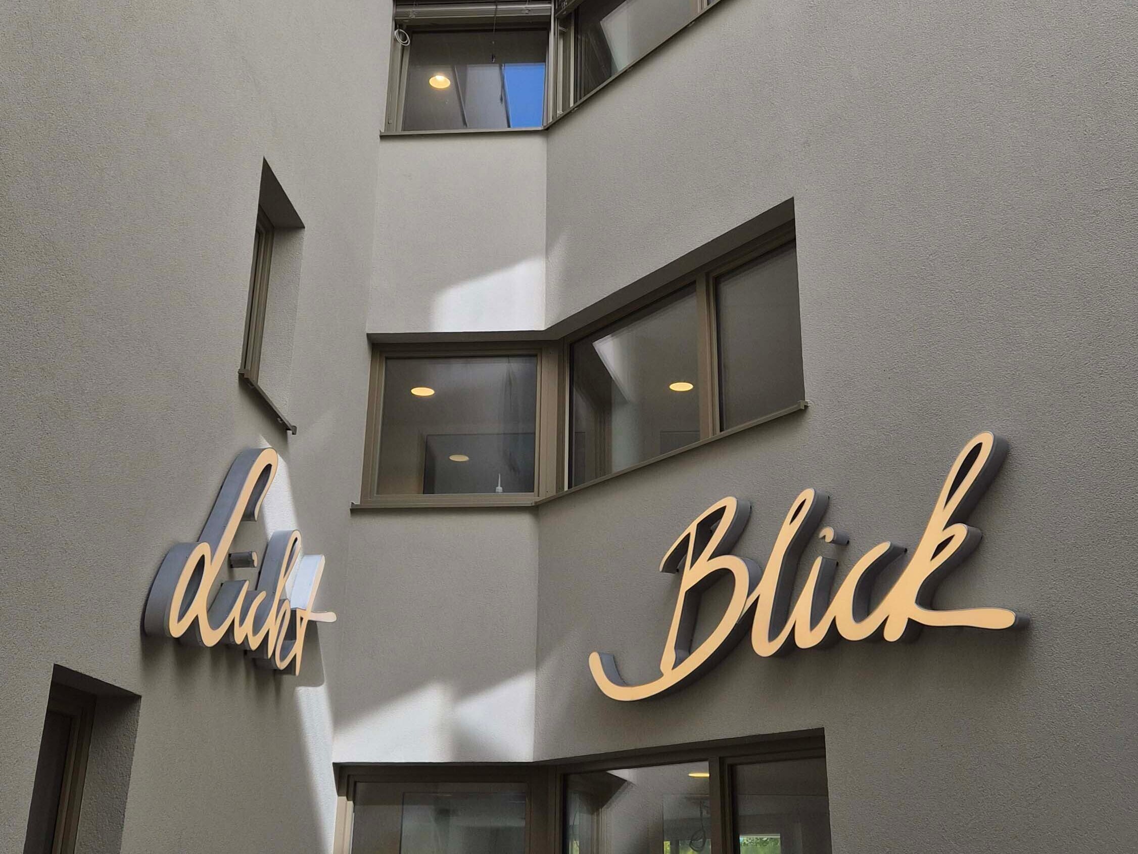 Innenhof eines modernen Gebäudes mit mehreren Fensterfronten. An den hellen Wänden sind in geschwungener Leuchtschrift die Wörter "Licht" und "Blick" angebracht.