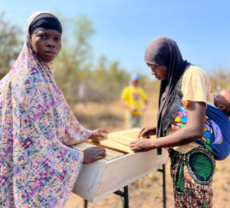 Two women are working outdoors on a modern wooden beehive. One of the women is looking toward the camera, while the other is standing sideways and carrying a small child on her back in a colorful wrap. In the background, a dry landscape typical of Burkina Faso can be seen, along with other participants.