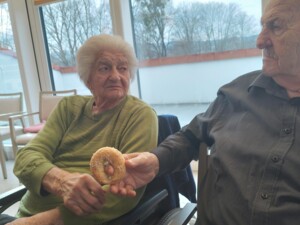 Eine ältere Bewohnerin mit weißem Haar sitzt im Rollstuhl und blickt zu einem Bewohner neben ihr, der einen kleinen Fastenbeugel in der Hand hält und zeigt. Beide sitzen in einem hellen Aufenthaltsraum mit großen Fenstern und schauen sich das Gebäck aufmerksam an.