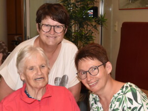 Drei Frauen - eine Seniorin und ihre beiden Angehörigen - sind auf dem Foto zusehen.  Die Seniorin sitzt links neben einer Angehörigen an einem Tisch. Die dritte Frau steht über den beiden lächelt über ihre Schultern in die Kamera. Im Hintergrund sind weitere Gäste in einem Gemeinschaftsraum zu sehen.